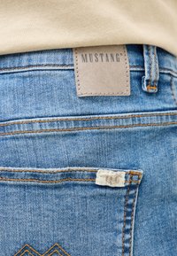 Šviesiai mėlynos džinsinės kelnės su matomu siuvinėjimu, rudos odos etiketė su užrašu "MUSTANG" ir balta etiketė ant galinio kišenės.