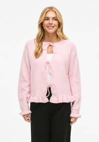 Rosa strikket cardigan med teksturert finish, som har volangkant, knyting og lange ermer. Brukt over en hvit topp og svarte bukser.