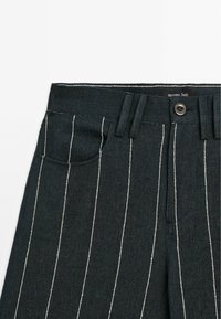 Pantalon en denim foncé avec des rayures verticales blanches, doté d'une fermeture par boutons, de passants pour ceinture et d'une poche avant. Design épuré et ajusté.