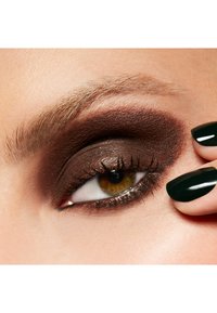 Gros plan sur un œil avec un fard à paupières brun foncé, des cils doux et un accent de vernis à ongles vert. La peau est lisse avec une texture légère.