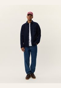 Navy blue button-up jacket with two front pockets, worn over a white shirt, paired with blue jeans and brown shoes, and a pink cap.