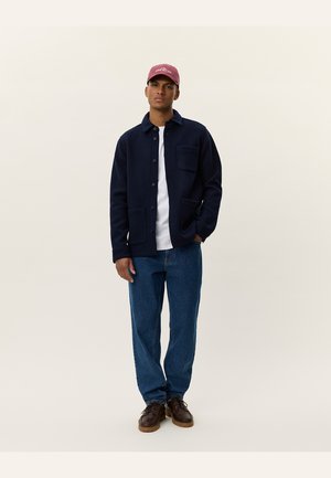 Navy blue button-up jacket with two front pockets, worn over a white shirt, paired with blue jeans and brown shoes, and a pink cap.