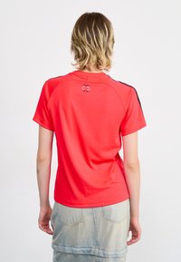 Raudona trumpa rankovė t-shirt su apvaliu kaklu, ant nugaros yra mažas logotipas, o pečių srityje – juodi akcentai. Lygus paviršius.