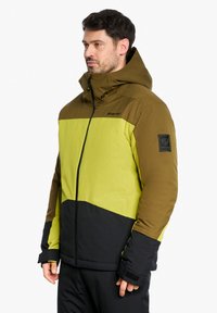 Giacca color block con torso giallo brillante, spalle verde oliva e parte inferiore nera, dotata di chiusura con zip e cappuccio regolabile.