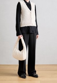 Person in schwarzem Hemd und Hose, mit einer cremefarbenen strukturierten, knopfartigen Weste, hält eine weiche cremefarbene Handtasche und steht auf einem Holzboden.