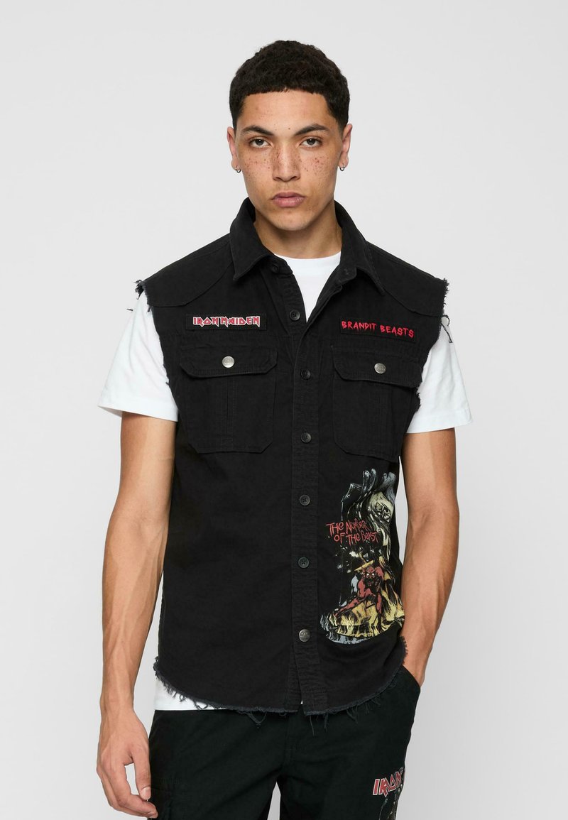 Gilet di denim nero con orli sfrangiati, due tasche frontali e stampa grafica. Presenta il logo "Iron Maiden" in rosso sul petto e sulla tasca.