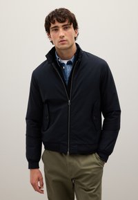 Giovane uomo che indossa una giacca blu navale con zip sopra una camicia blu a bottone e pantaloni verde oliva, in piedi contro uno sfondo chiaro e senza fronzoli.
