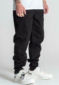 Pantalones de mezclilla negros con un corte relajado y piernas ligeramente ajustadas, combinados con zapatillas blancas que presentan detalles de marca y cordones texturizados.