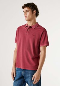 Camisa polo de algodón roja con cuello, dos botones y mangas cortas. Presenta un pequeño logo en el pecho. Llevada con jeans de mezclilla azules.