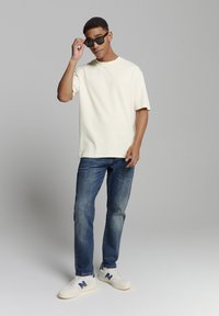 Ecru kleurig oversized katoenen t-shirt, blauwe denim jeans en witte sneakers met marineblauwe accenten. Persoon past zwarte zonnebril aan.