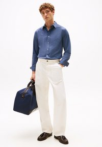 Langarm blauw geruit overhemd, witte relaxed-fit broek en donkerbruine instappers. Houdt een marineblauwe sporttas met donkere banden vast.