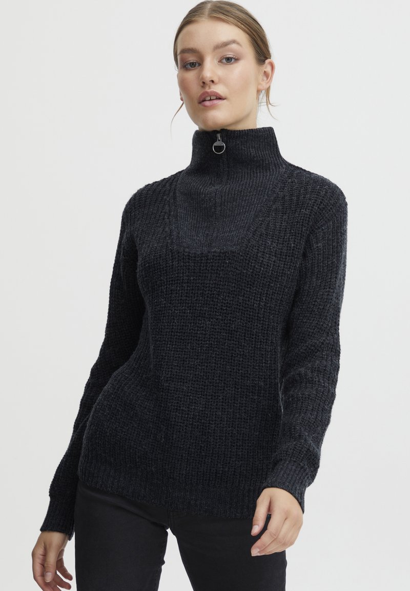 Pull en maille noir avec un col montant, présentant une fermeture éclair quart à la nuque, un tissu texturé et des manches ajustées.