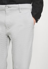 Lichtgrijze denim broek met een gladde textuur, zakken aan de voorkant en een knoopsluiting in de taille. Gecombineerd met een zwart overhemd met knopen.