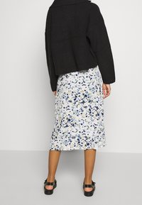 Un pull court en tricot noir à côtes porté par-dessus une jupe midi fleurie aux motifs bleus, jaunes et verts. Des sandales noires à semelle épaisse.