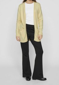 Gul stickad kofta med öppen front och två fickor, ihop med en vit topp och svarta utsvängda byxor. Slät textur, avslappnad passform.