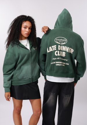 Frau und Mann tragen dunkelgrüne Kapuzenpullover; die Frau blickt nach vorne, der Mann zeigt den Rücken mit dem Schriftzug "Late Dinner Club" auf dem Hoodie.