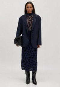 Marineblazer über einem transparenten, floralen Spitzentop, kombiniert mit einem Rock mit Blumendruck und schwarzen kniehohen Stiefeln mit Absatz. Schwarze Handtasche in der Hand.