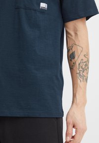 Navy blue cotton t-shirt with short sleeves, featuring a chest pocket and a fabric label; worn with black pants. Arm tattoo visible.