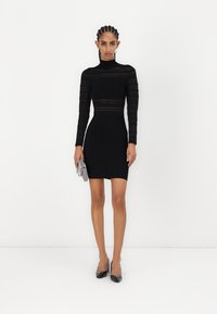 Svart langermet tettsittende kjole med transparente striper, høy hals og minikjolelengde. Modellen holder en sølvfarget metallisk clutch.