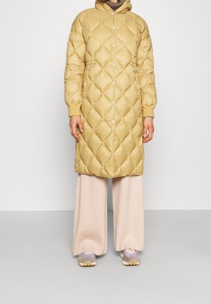 Person, die einen langen, beige gesteppten Mantel mit Druckknöpfen, cremefarbene Hosen mit weitem Bein und hellviolette Turnschuhe trägt und vor einem schlichten Hintergrund steht.