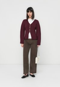 Vrouw staat tegen een witte achtergrond, draagt een bordeauxrode cardigan met knopen over een witte top, bruine wijde broek, zwarte flats en houdt een kleine witte tas vast.