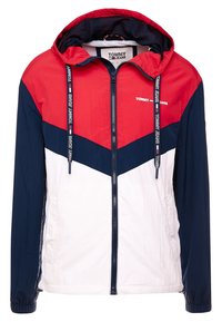 Color-blocked hooded jacket in red, navy, and white. Features a zip closure, elastic cuffs, and a drawstring hood. "TOMMY JEANS" logo on chest.