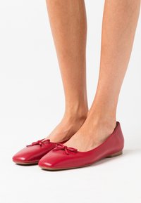 Des ballerines en cuir rouge présentent une finition lisse, un bout arrondi et des nœuds décoratifs à l'avant. La semelle est plate et flexible.