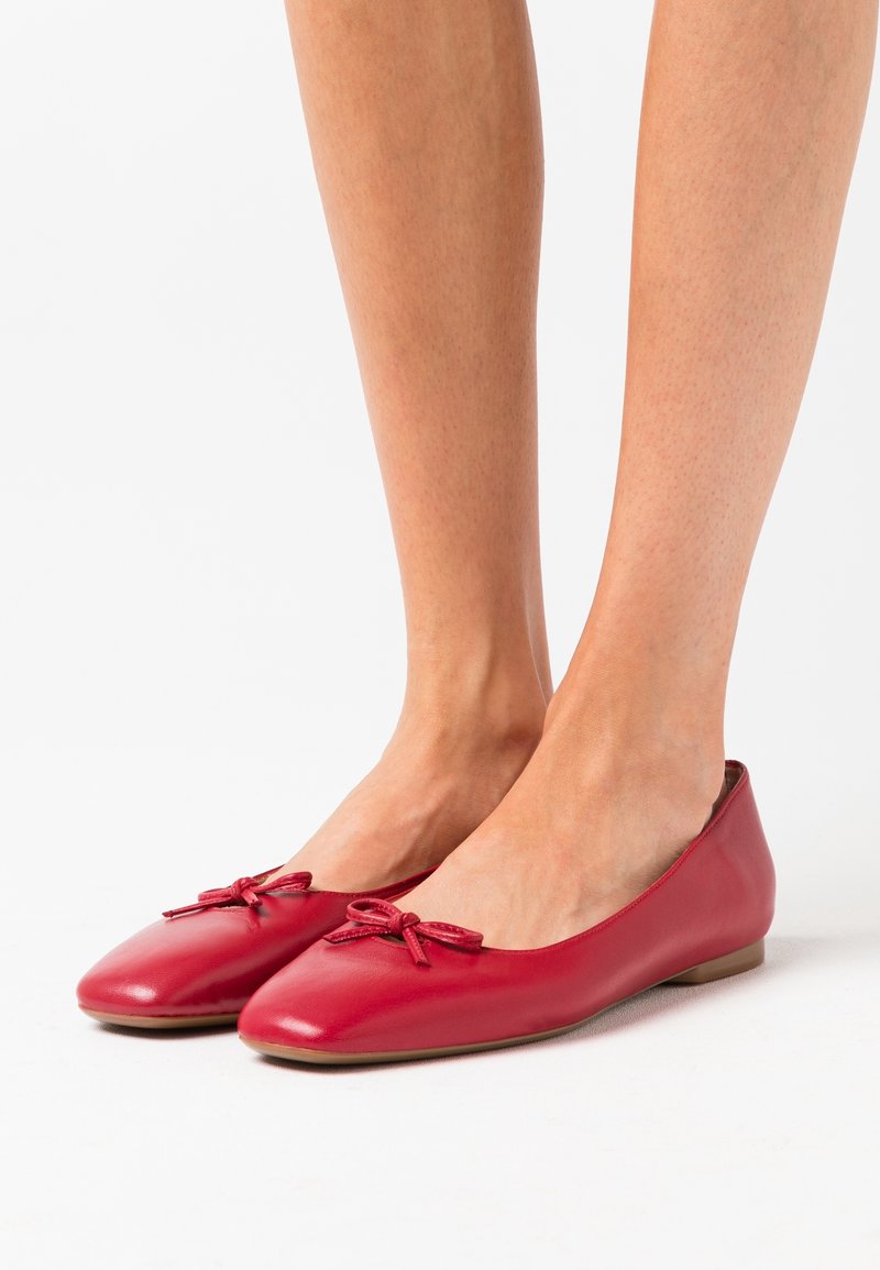 Des ballerines en cuir rouge présentent une finition lisse, un bout arrondi et des nœuds décoratifs à l'avant. La semelle est plate et flexible.