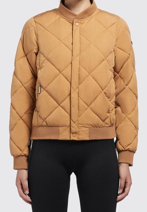 Femme portant une veste aviateur matelassée beige avec col, poignets et ourlet côtelés, associée à un pantalon noir, vue de face, visage non visible.