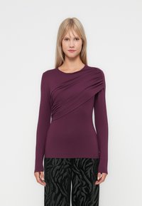 Haut à manches longues en violet profond avec un détail drapé à l'avant ; texture lisse et coupe ajustée, associé à un pantalon noir à motifs.