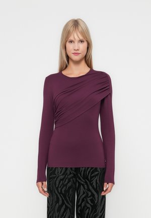 Top a maniche lunghe di un profondo viola con dettaglio drappeggiato sul davanti; texture liscia e design aderente, abbinato a pantaloni neri a fantasia.