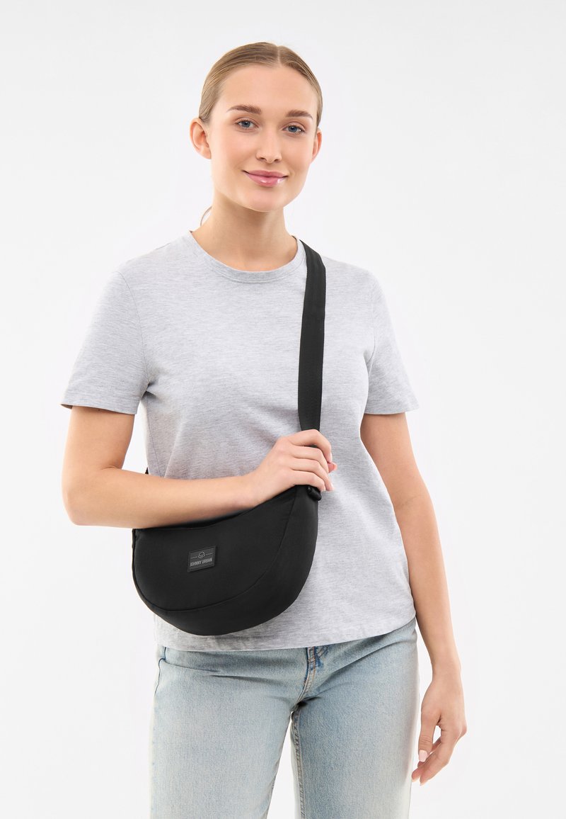 Czarna torba crossbody z zaokrąglonym kształtem, regulowanym paskiem i dyskretną łatą z logo; gładka tekstura i minimalistyczny design.