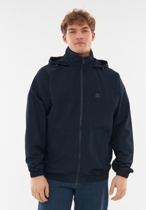 Marineblå vindjakke med hætte, lynlås foran, elastiske ærmer og et patch-logo på brystet. Letvægtsstof med en glat tekstur.