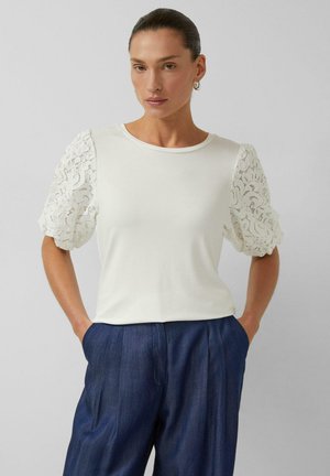 Frau mit nach hinten geglättetem Haar, die ein weißes Oberteil mit Spitzen-Puffärmeln und hoch taillierte, lockere dunkelblaue Hose trägt, Hände in den Taschen.