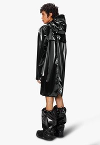 Gabardina negra, brillante y impermeable con capucha y un detalle de volantes en la parte trasera, combinada con botas impermeables negras y arrugadas.