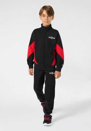 Ragazzo giovane che cammina in avanti indossando una giacca a zip nera e rossa e pantaloni abbinati, con sneakers nere dotate di lacci rossi e suole bianche.