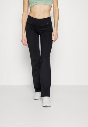 Kvinde iført sorte Nike leggings med høj talje, lysegrøn sports-bh og hvide sneakers, stående mod en ensfarvet baggrund.