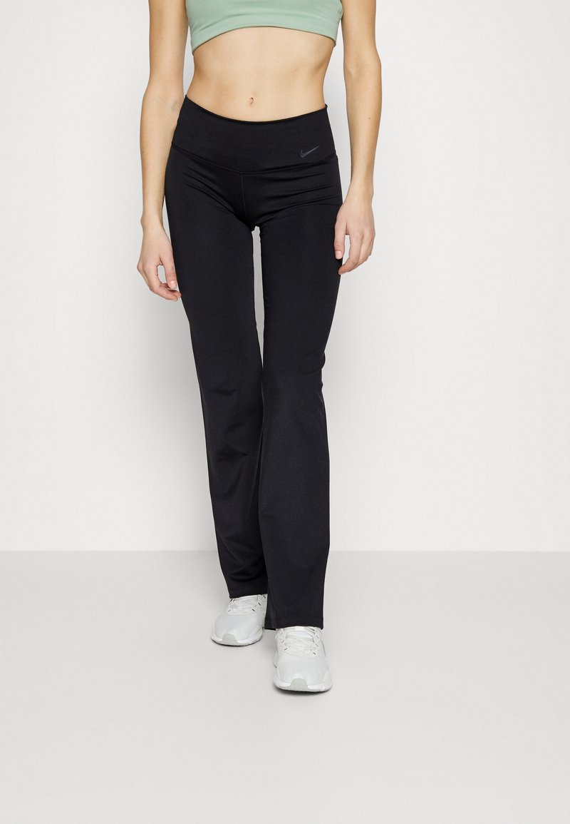 Femme portant des leggings noirs taille haute Nike, un soutien-gorge de sport vert clair et des baskets blanches, debout devant un fond uni.