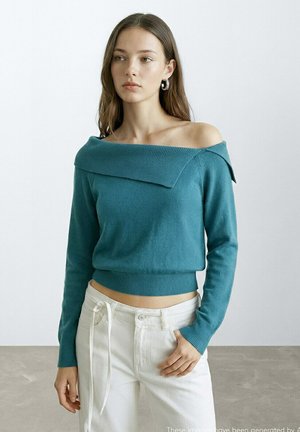 Giovane donna che indossa un maglione scivolato sulle spalle color teal e pantaloni bianchi, in piedi con una mano in tasca contro uno sfondo semplice.