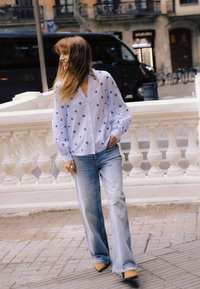 Donna con capelli lunghi in piedi vicino a una balaustra bianca, indossa una camicetta bianca fantasia, jeans a gamba larga e scarpe gialle con punta, su un marciapiede cittadino.