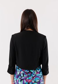 Giacca nera corta con maniche a tre quarti, tessuto liscio, design semplice, indossata sopra una gonna a fiori multicolore.