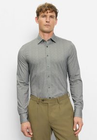 Ein Mann steht vor einer weißen Wand, trägt ein eng anliegendes, graues, gemustertes Hemd mit Knöpfen und olivgrüne Hosen und blickt mit neutralem Gesichtsausdruck nach vorne.