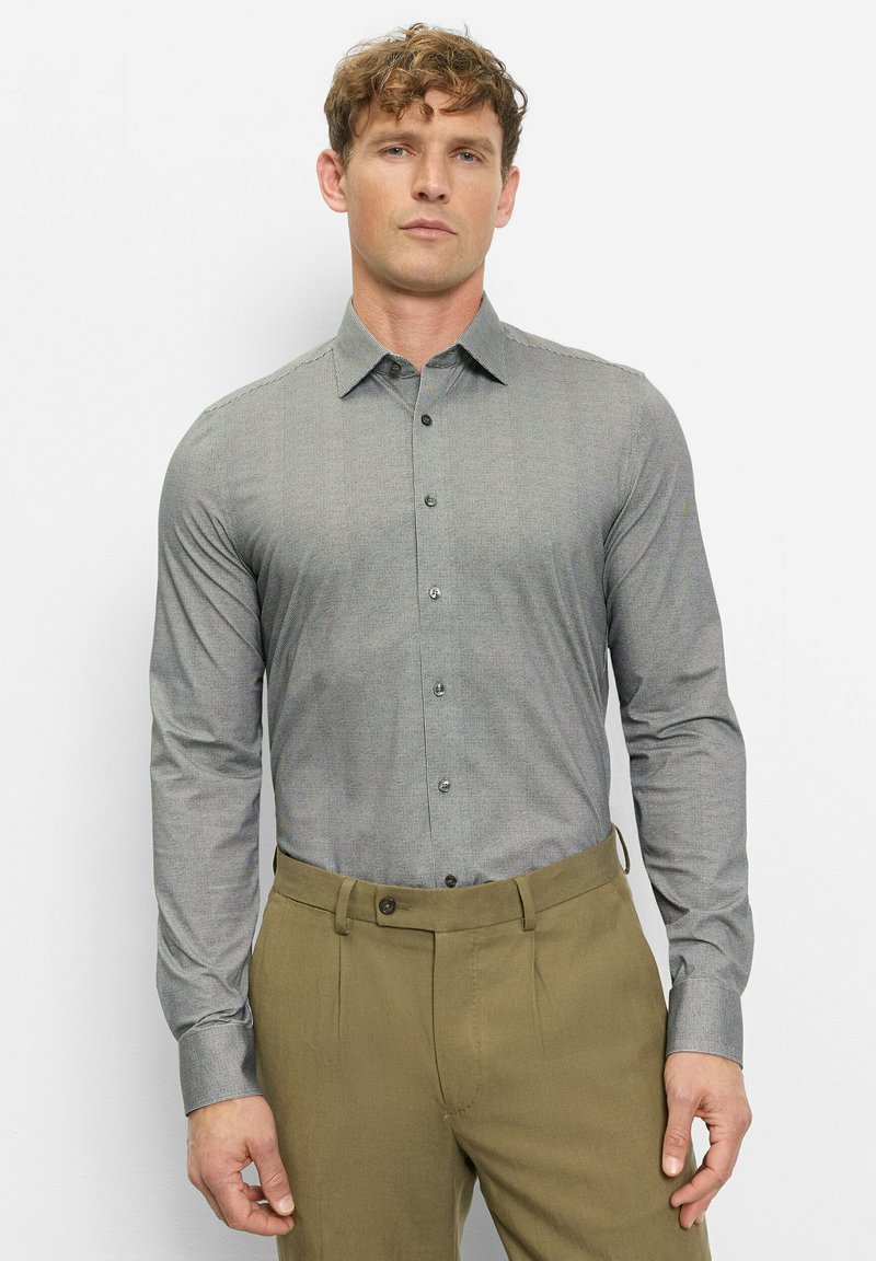 Ein Mann steht vor einer weißen Wand, trägt ein eng anliegendes, graues, gemustertes Hemd mit Knöpfen und olivgrüne Hosen und blickt mit neutralem Gesichtsausdruck nach vorne.