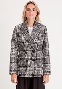 Double-breasted jacket in black and white houndstooth pattern, featuring a notched collar, long sleeves, and black buttons. Textured fabric.