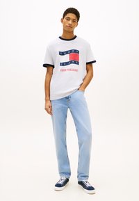 Ung mann står med den ene hånden i lomma, iført hvit Tommy Jeans t-skjorte, lyseblå jeans og marineblå sneakers mot ensfarget bakgrunn.