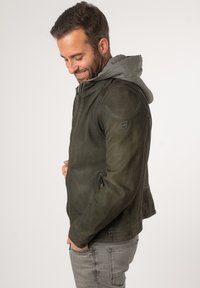 Dunkelgrüne Lederjacke mit Reißverschluss vorne, zwei seitlichen Reißverschlüssen und grauem Kapuzeneinsatz. Glatte Textur, taillierte Passform und Logodetail an der Schulter.