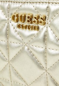 Superficie de cuero acolchado dorado con el logotipo metálico dorado "GUESS EST 1981" embossado en ella.
