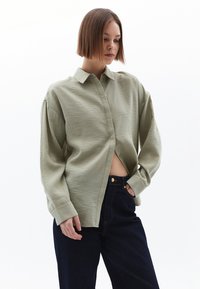 Camicia button-up di un verde chiaro, con texture, colletto, maniche lunghe e vestibilità rilassata, abbinata a pantaloni di denim blu scuro.