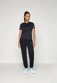 Čierne tričko s krátkym rukávom s logom na rukáve, spojené s čiernymi teplákmi s malým ružovým logom. Model má na sebe biele tenisky.