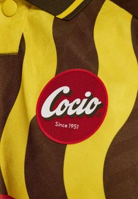 Żółto-brązowy materiał w wzory z okrąłą czerwoną łatką z logo "Cocio" i tekstem "Since 1951". Teksturowana powierzchnia, widoczny guzik.